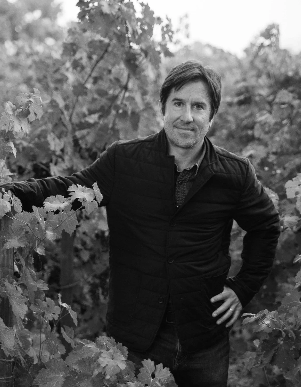 A man standing in a vineyard, smiling, with grapevine leaves surrounding him, in black and white.