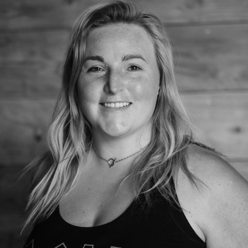 A black and white portrait of a smiling young woman with long, wavy blonde hair, wearing a sleeveless top and a necklace, standing against a wooden background.