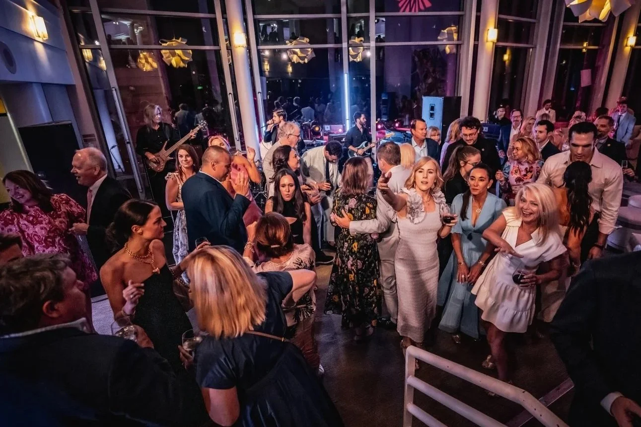 People dancing and socializing at a lively party or celebration in an indoor venue with large glass windows, with musicians performing live music in the background.