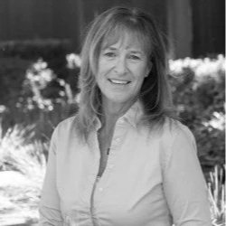 A woman smiling outdoors with flowers and greenery in the background.
