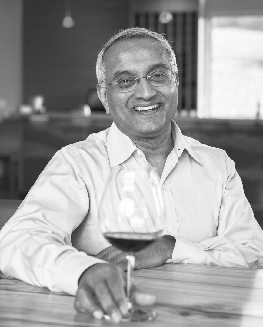 A smiling man wearing glasses and a white shirt, sitting at a table with a glass of red wine in front of him, in a well-lit indoor setting.