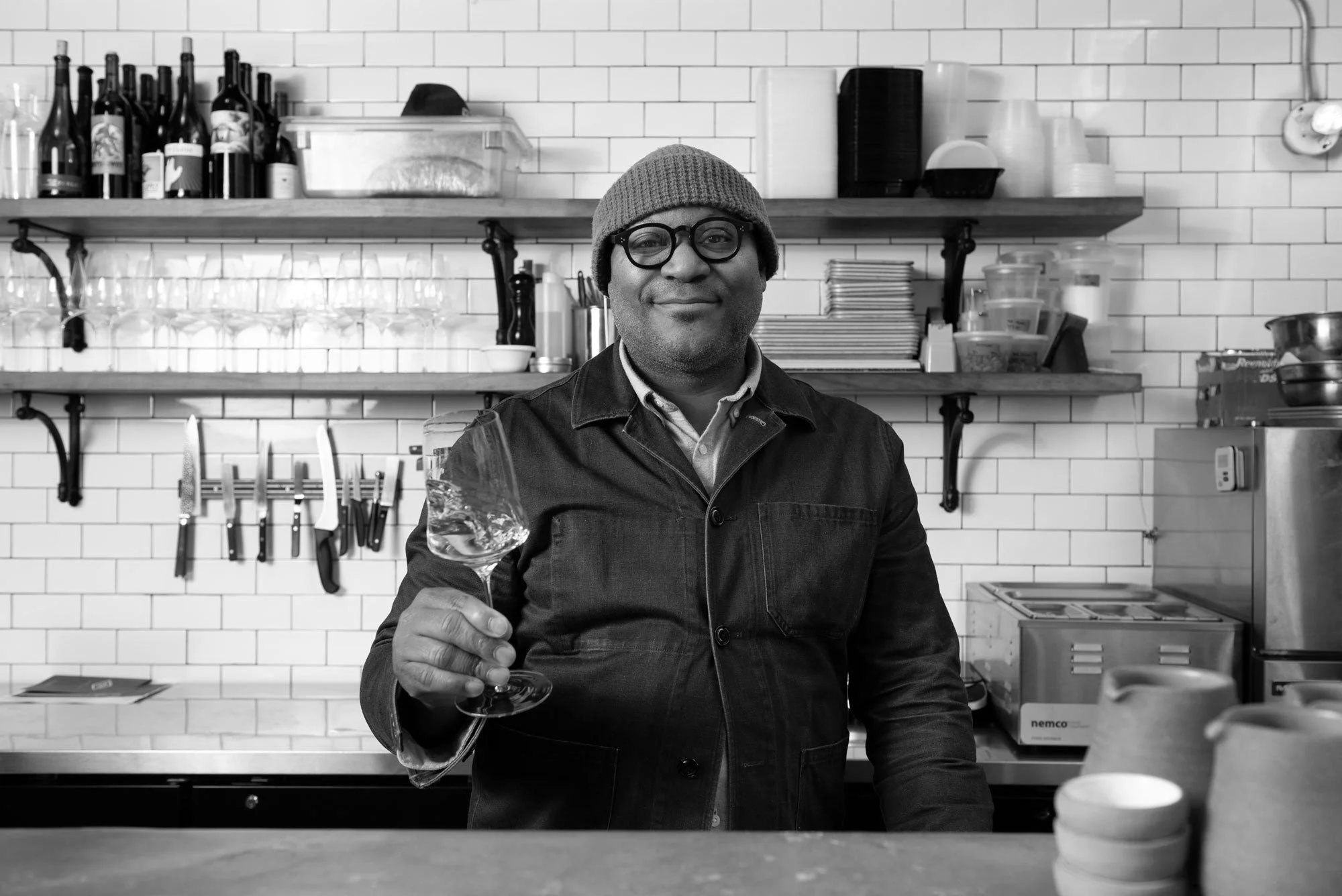 A man in a beanie and glasses holding a wine glass in a restaurant or cafe kitchen with shelves of bottles, glasses, and kitchen tools in the background.