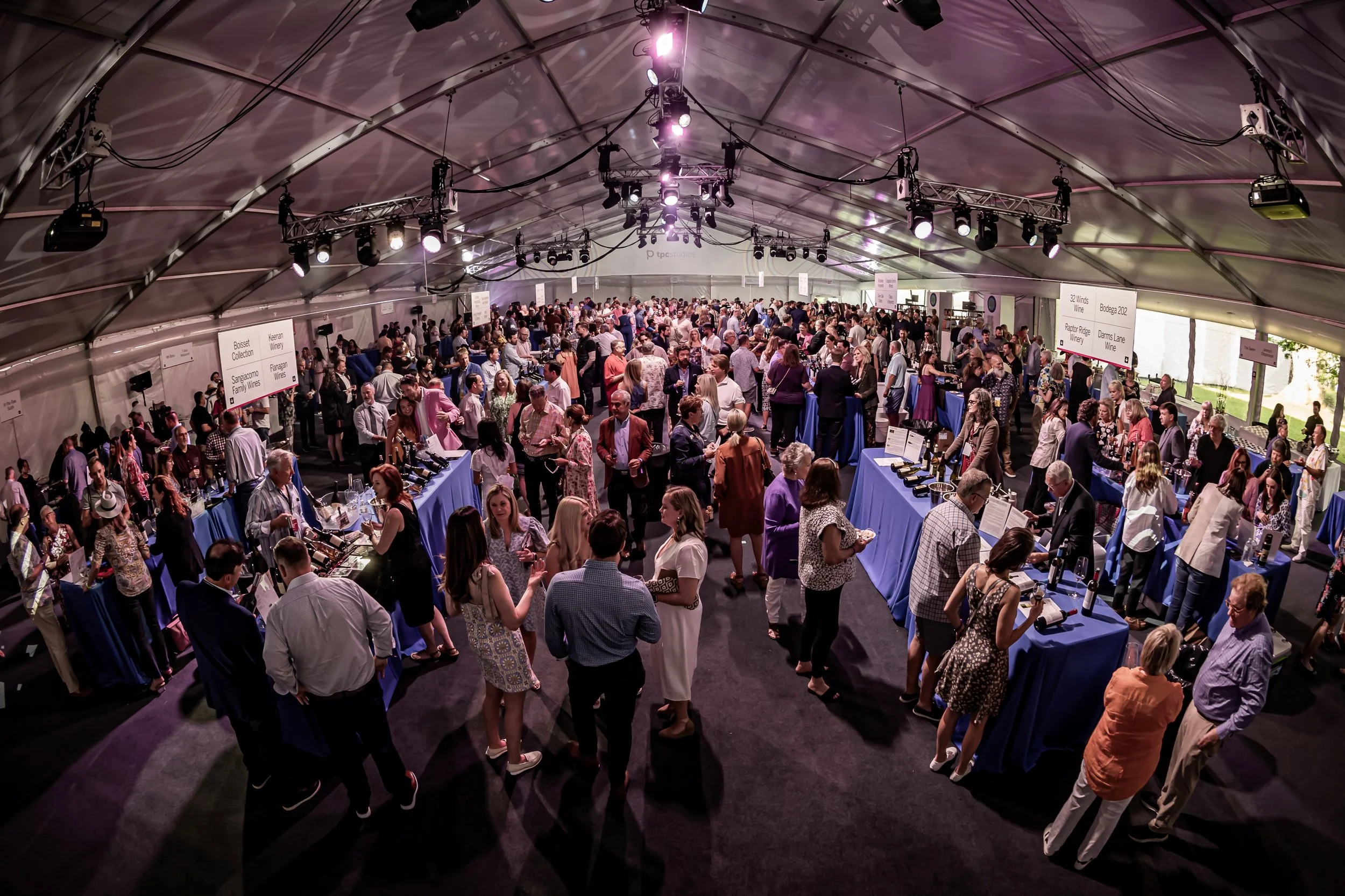 Large indoor wine tasting event with many attendees browsing tables of wine bottles, under a curved ceiling with lighting and signage.