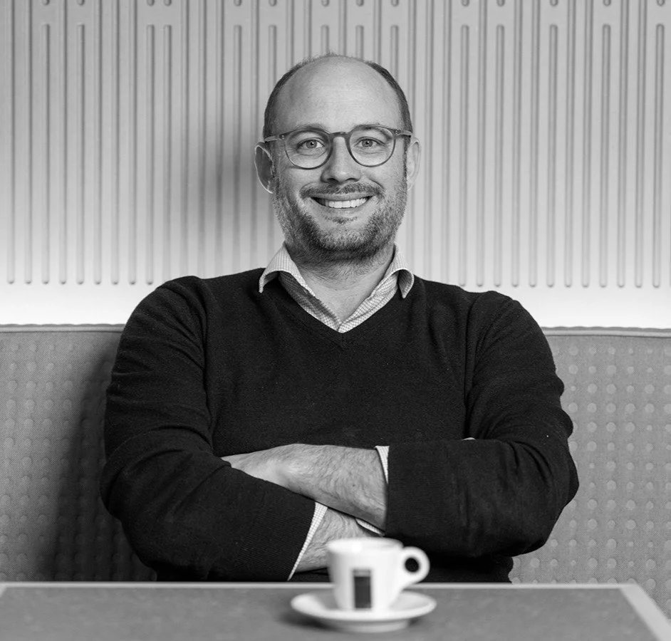 Black and white photo of a man with glasses smiling, seated with arms crossed, in a cafe or restaurant, with a small cup of coffee on the table in front of him.