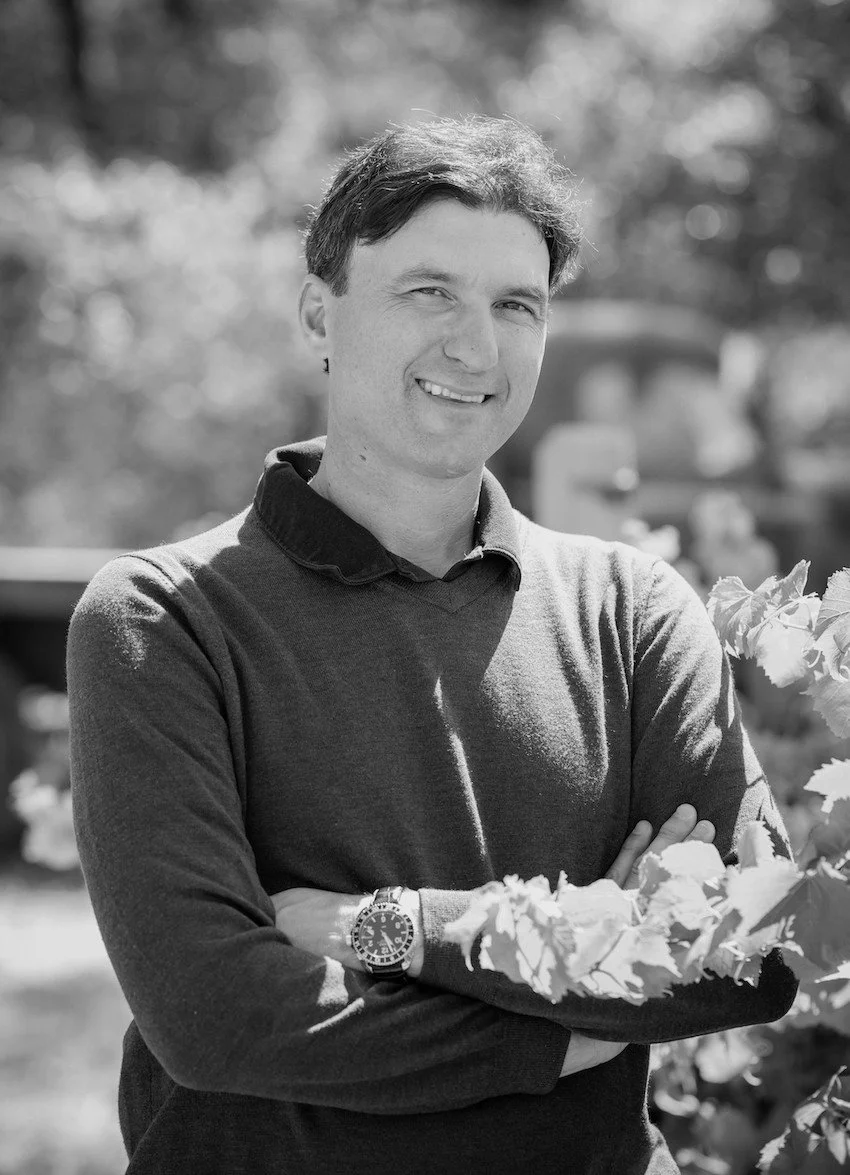 A smiling man standing outdoors with arms crossed, wearing a sweater and wristwatch, in a natural setting with trees and blurred vehicles in the background.
