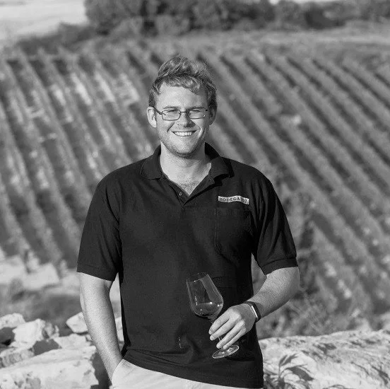 Man smiling, holding a wine glass outdoors in a vineyard.