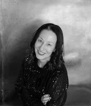 Black and white photo of a smiling woman with dark, wavy hair, wearing a dark, sparkling top, standing against a plain background.