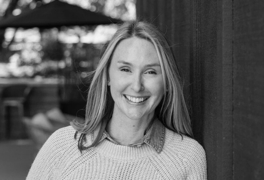 Black and white photo of a smiling woman with long hair, leaning against a wooden wall, outdoors with some blurred trees and furniture in the background.