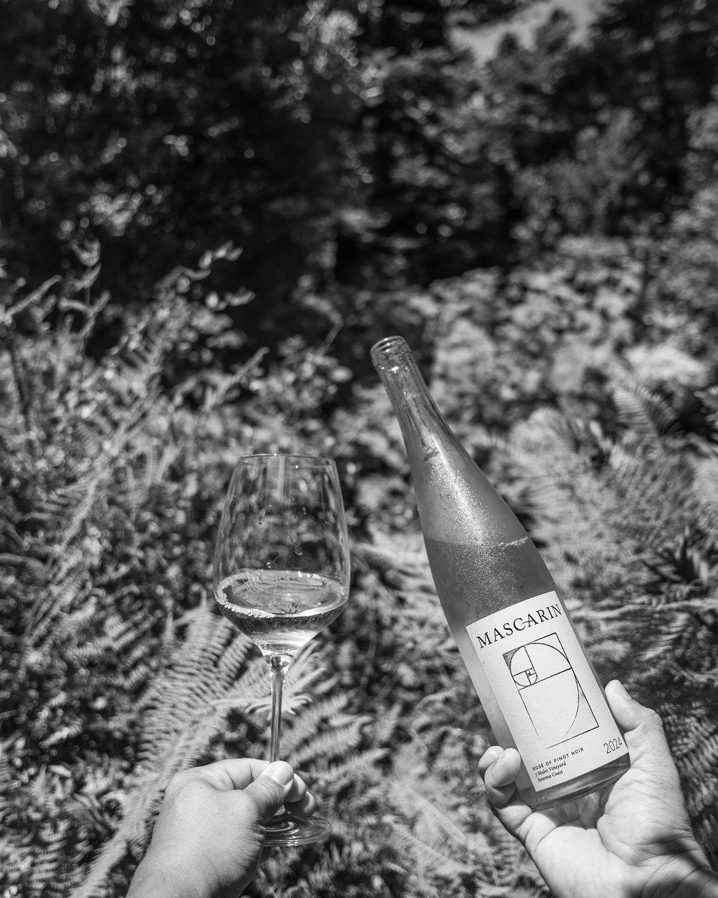 A person holding a glass of wine in one hand and a bottle of Mascarin rosé wine in the other outdoors, with dense ferns in the background.