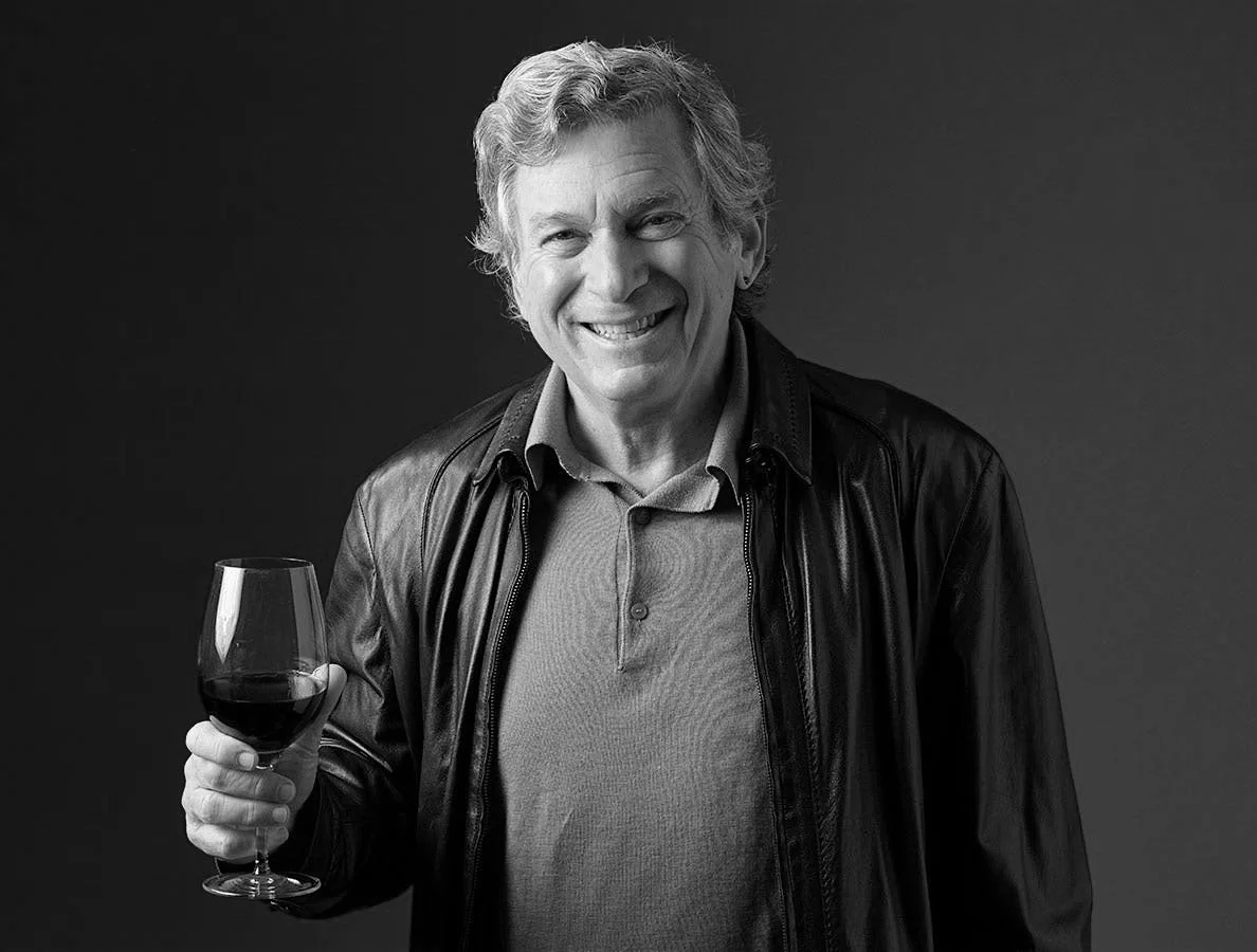 A smiling elderly man with gray hair holding a glass of red wine, wearing a leather jacket over a polo shirt, standing against a plain dark background.