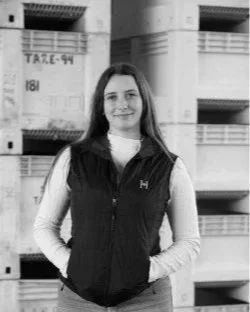 Young woman with long hair smiling, wearing a black vest over a white long-sleeve shirt, standing outdoors near residential buildings.
