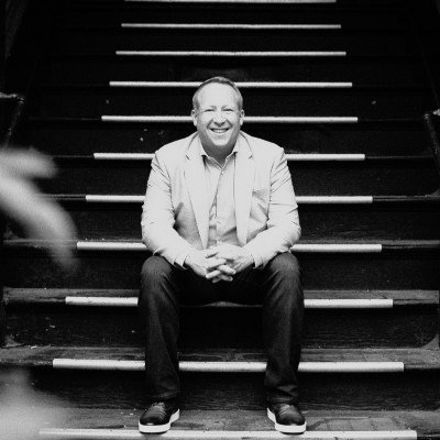 A man sitting on stairs, smiling, in a casual suit and sneakers.