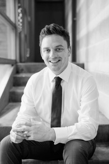 A smiling man in a white shirt and dark tie sitting on stairs near a window in a professional setting.