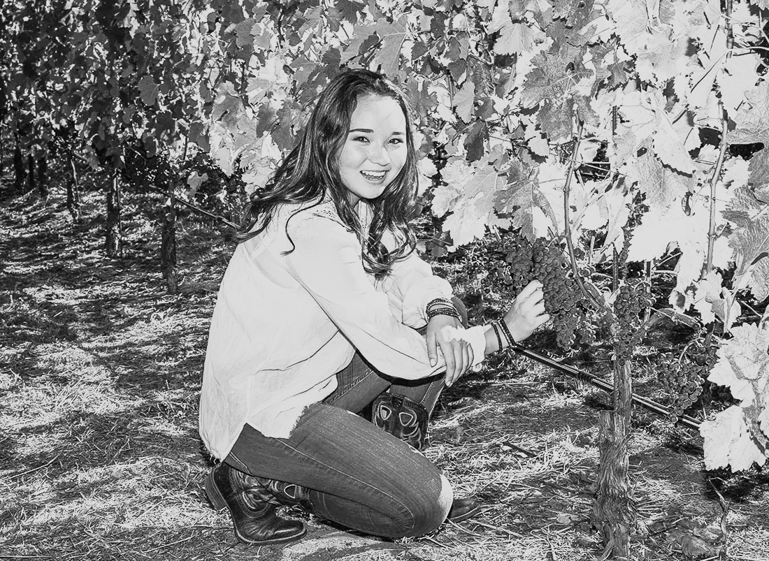 A young woman with long hair and a smile, wearing a light-colored jacket and dark jeans, kneeling in a vineyard, harvesting grapes from a vine.