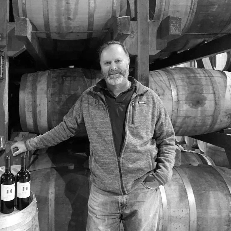 A man with a beard and short hair stands in a distillery, surrounded by stacked wooden barrels, with some wine bottles on a barrel in front of him.
