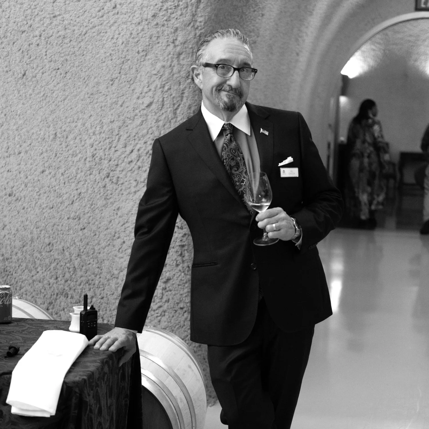 A man in a black suit, glasses, and tie holding a wine glass, standing next to a table with a napkin and a walkie-talkie, in a hallway with an arched ceiling and other people in the background.