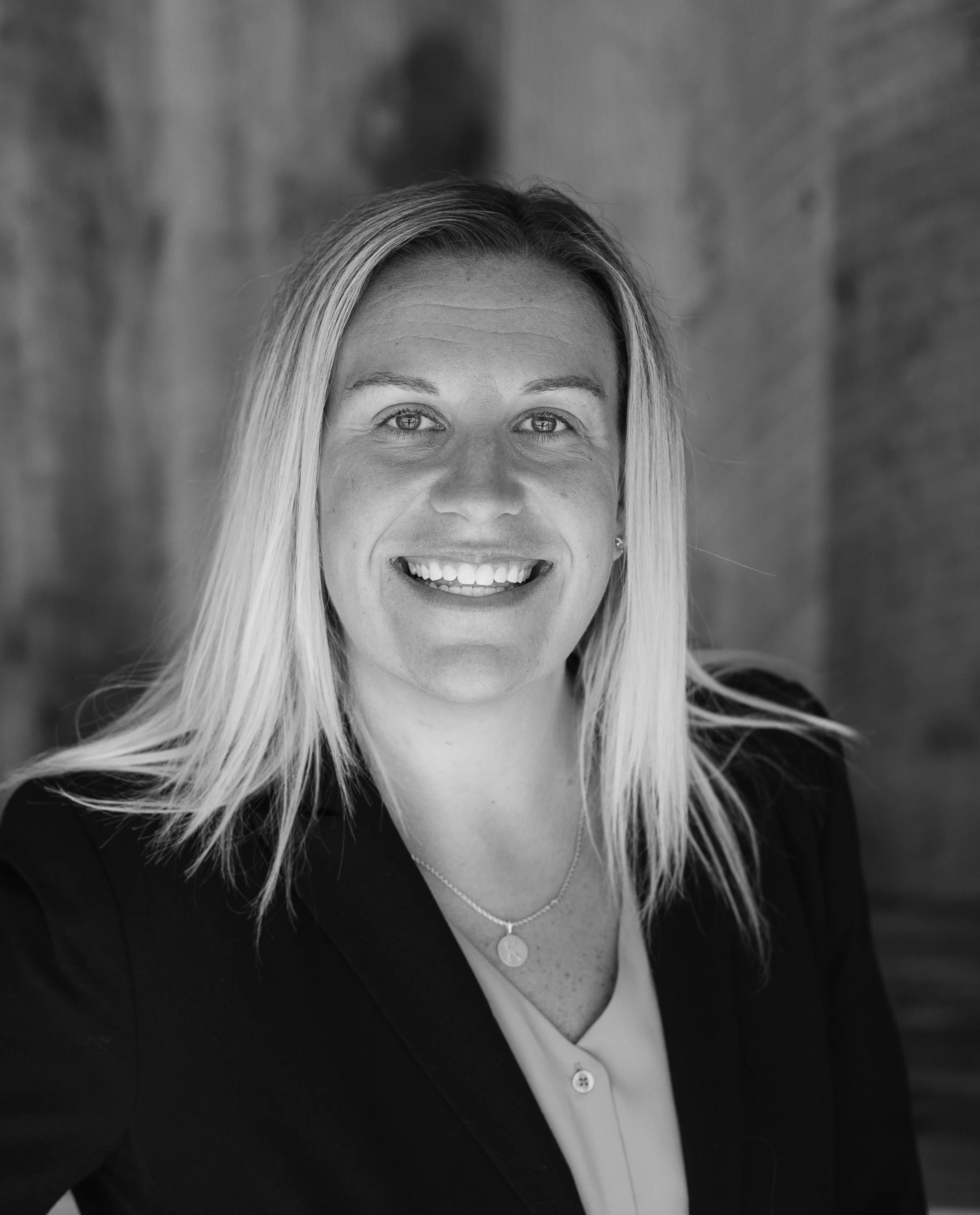 A woman with blonde hair smiling, wearing a dark blazer, light blouse, and a necklace, in front of a textured background.