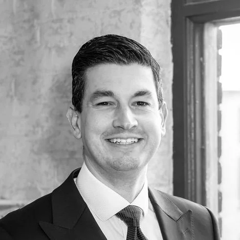 Black and white portrait of a smiling man in a suit and tie, standing near a window with a brick wall background.