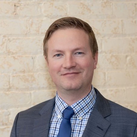 portrait of a man with light brown hair, wearing a gray suit, blue checkered shirt, and blue tie, standing in front of a beige brick wall.