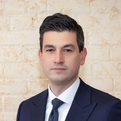 A professional man with dark hair in a suit and tie, standing against a light-colored brick wall.