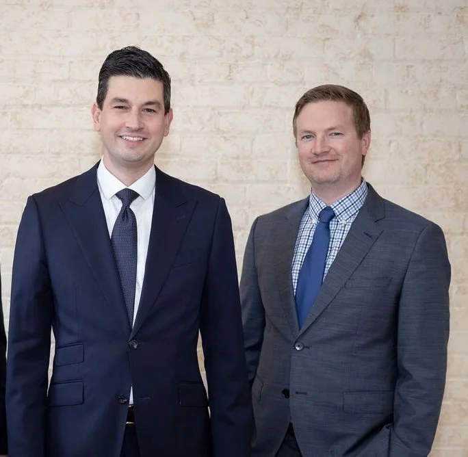 Two professionally dressed men stand side by side smiling in front of a beige brick wall.