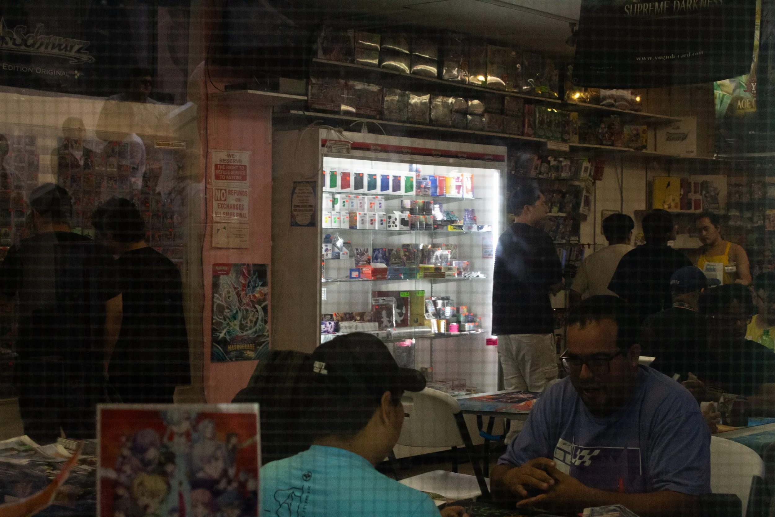 An illuminated case full of colorful prizes shines in a darkly lit store. Customers are seated or are standing around the case.