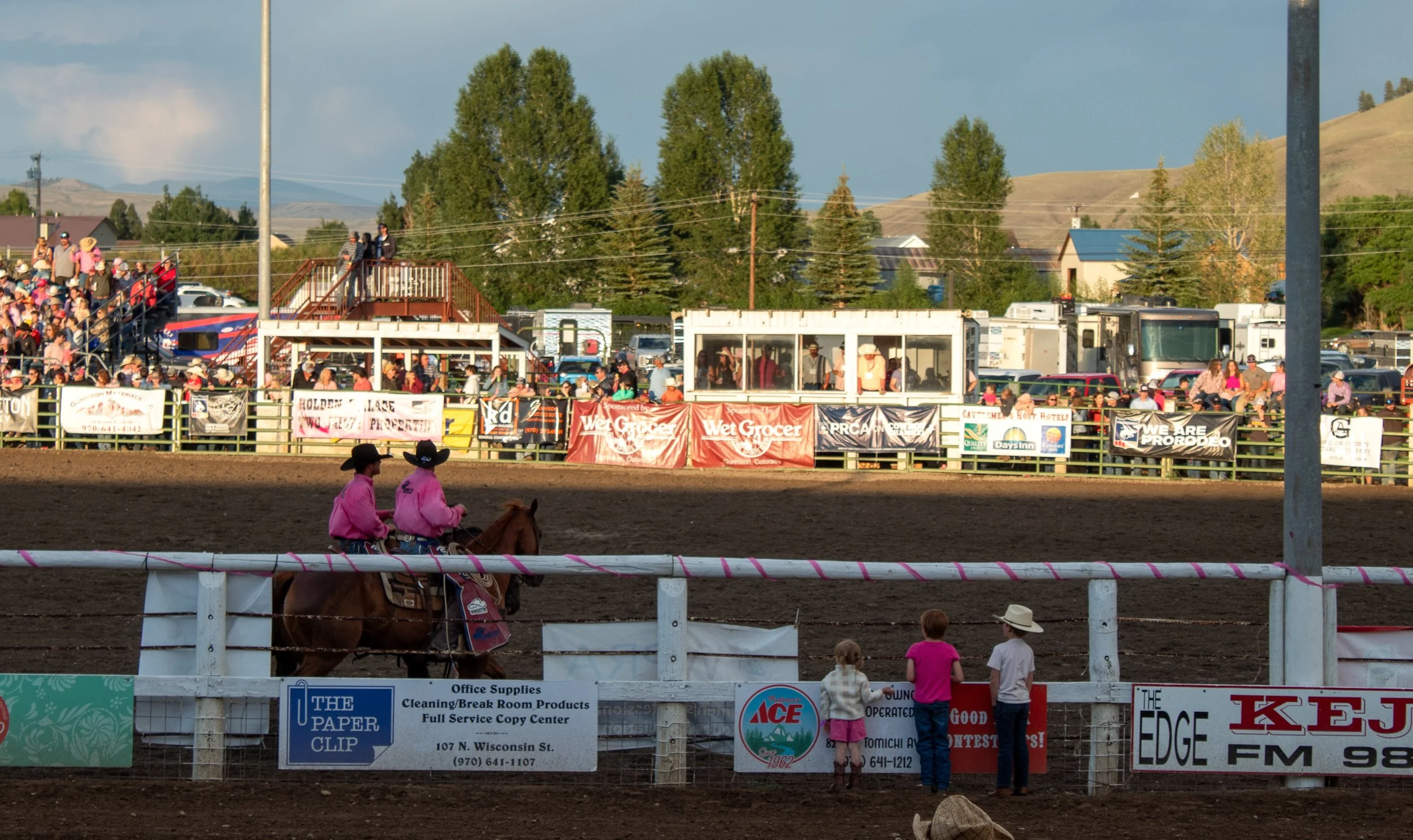 Two cowgirls sit next to each other atop their horses at a rodeo. Three small children stare at them from the fence. The fence is draped with ads for local businesses and a crowd stands in the bleaches in the background.