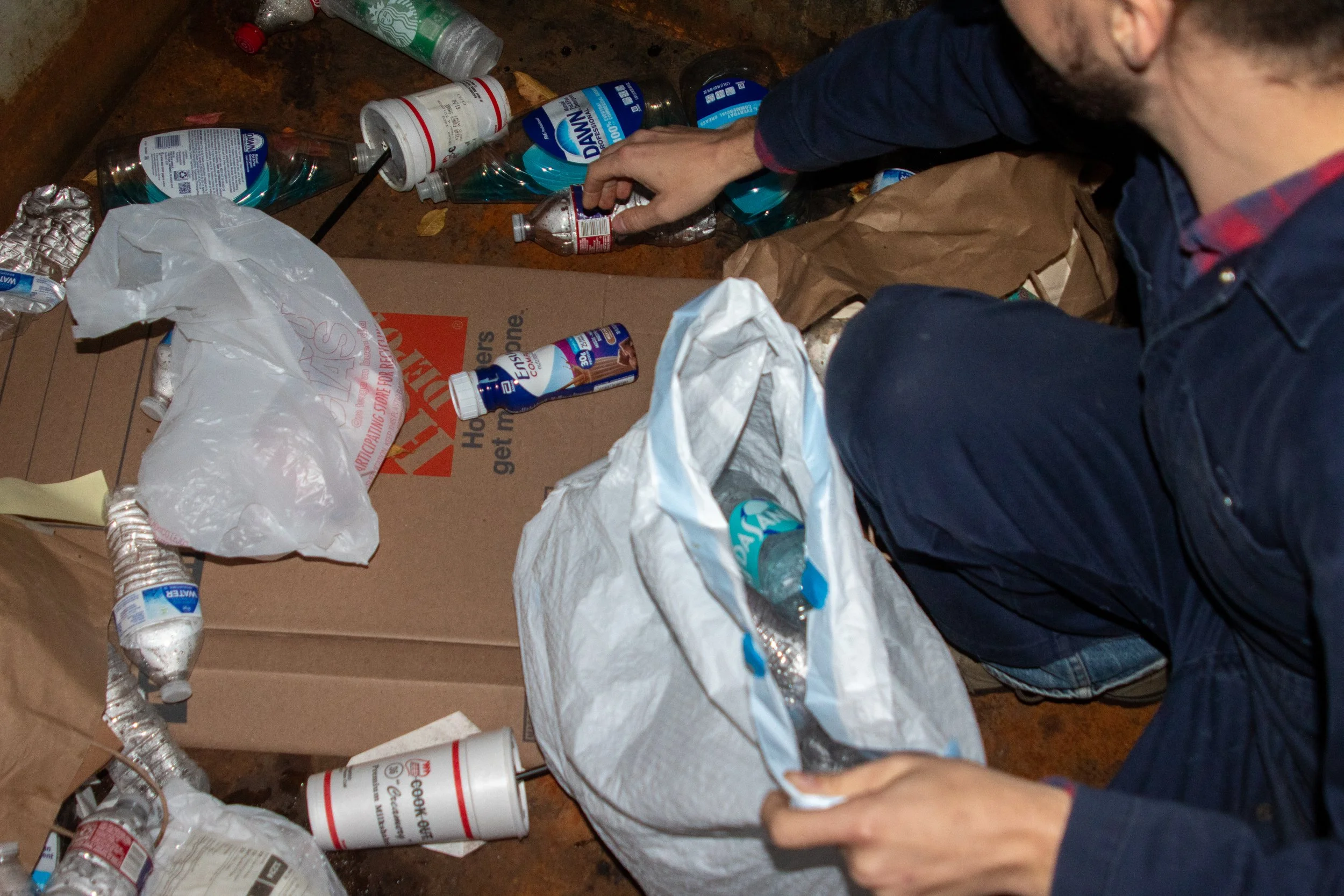 Scattered on the floor are to-go cups, empty protein shake bottles, water bottles, and Dawn dish soap bottles partly full. A Home Depot cardboard box rests beneath the trash pile. A man reaches to put the trash into his trash bag.