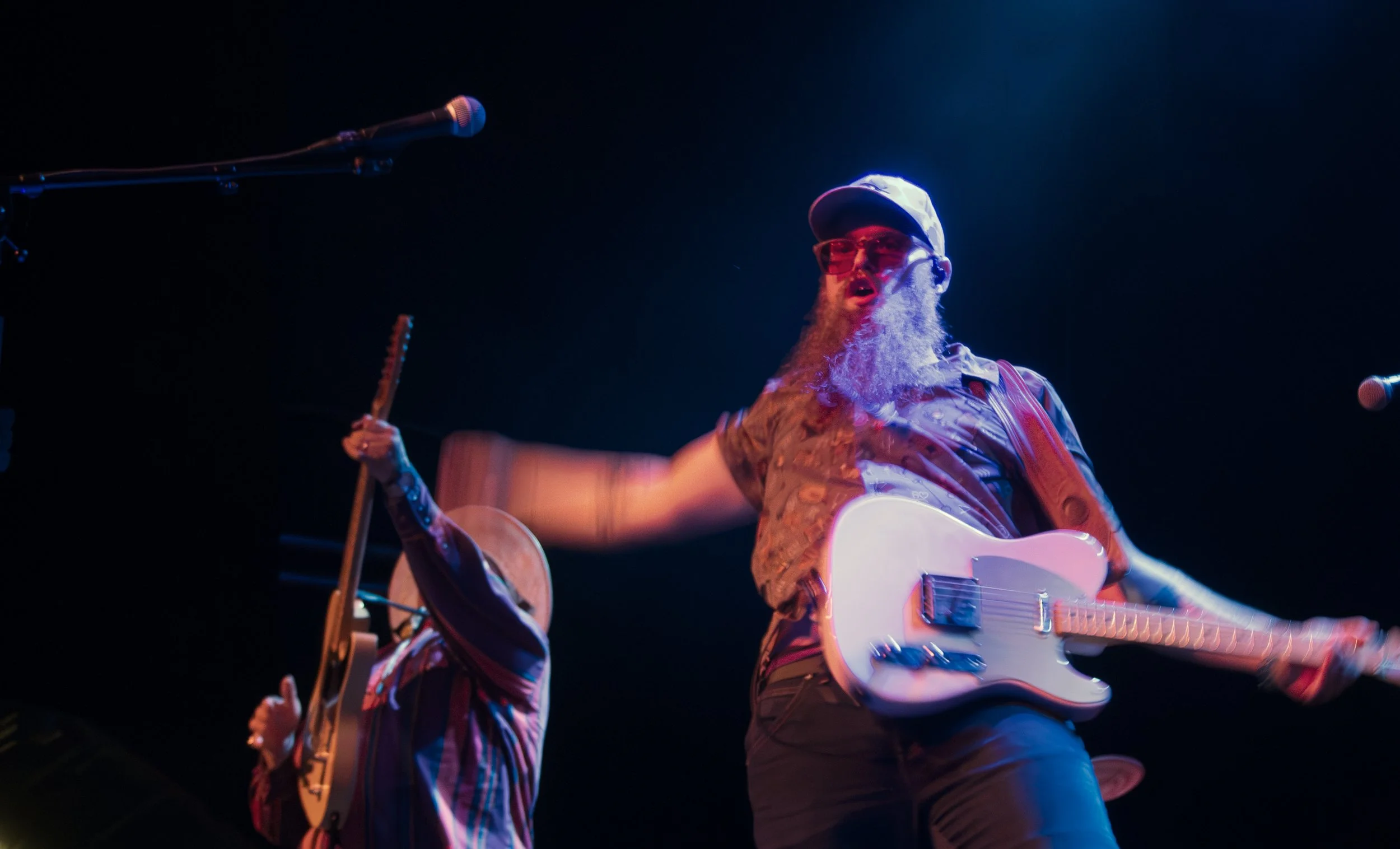 A man and another person play electric guitar on stage. The background is dark while the stagelights shine on them. The man's arms are blurred as he plays the gutiar.