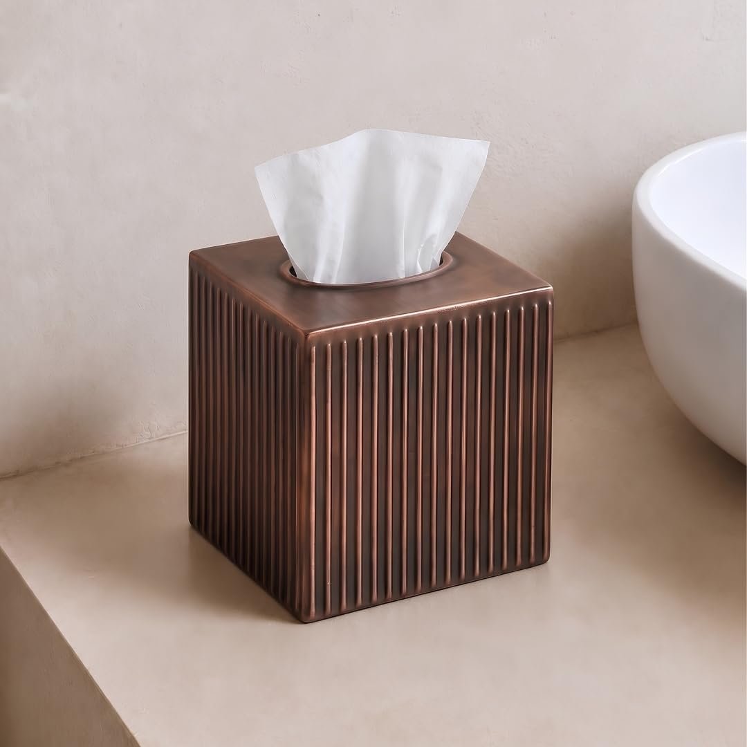 Rectangular copper-colored tissue box with vertically ridged design and a tissue protruding from the top, placed on a beige surface next to a white ceramic basin.