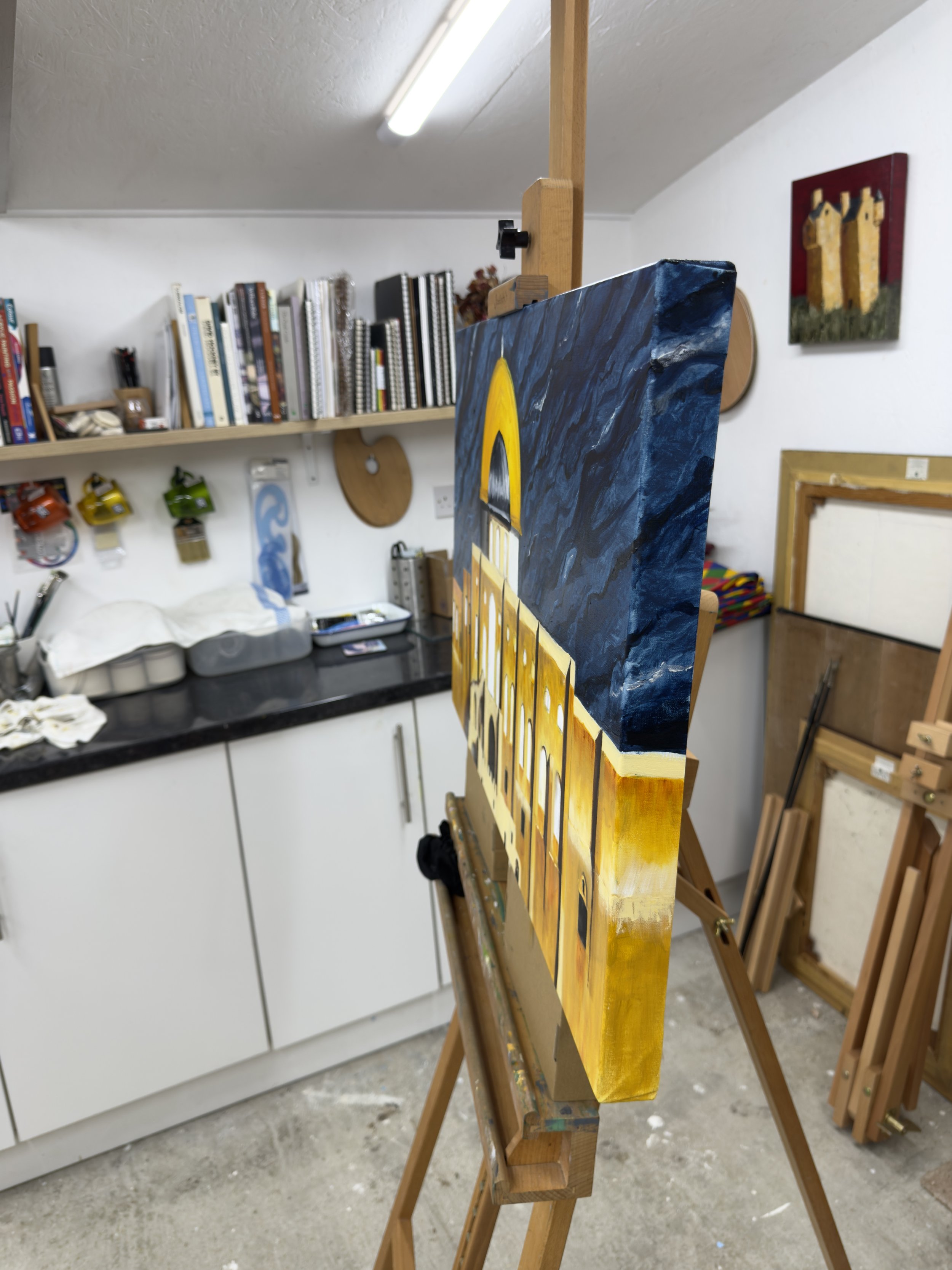An artist's studio with a partially completed painting on an easel, features a building with a yellow facade and a blue sky background.