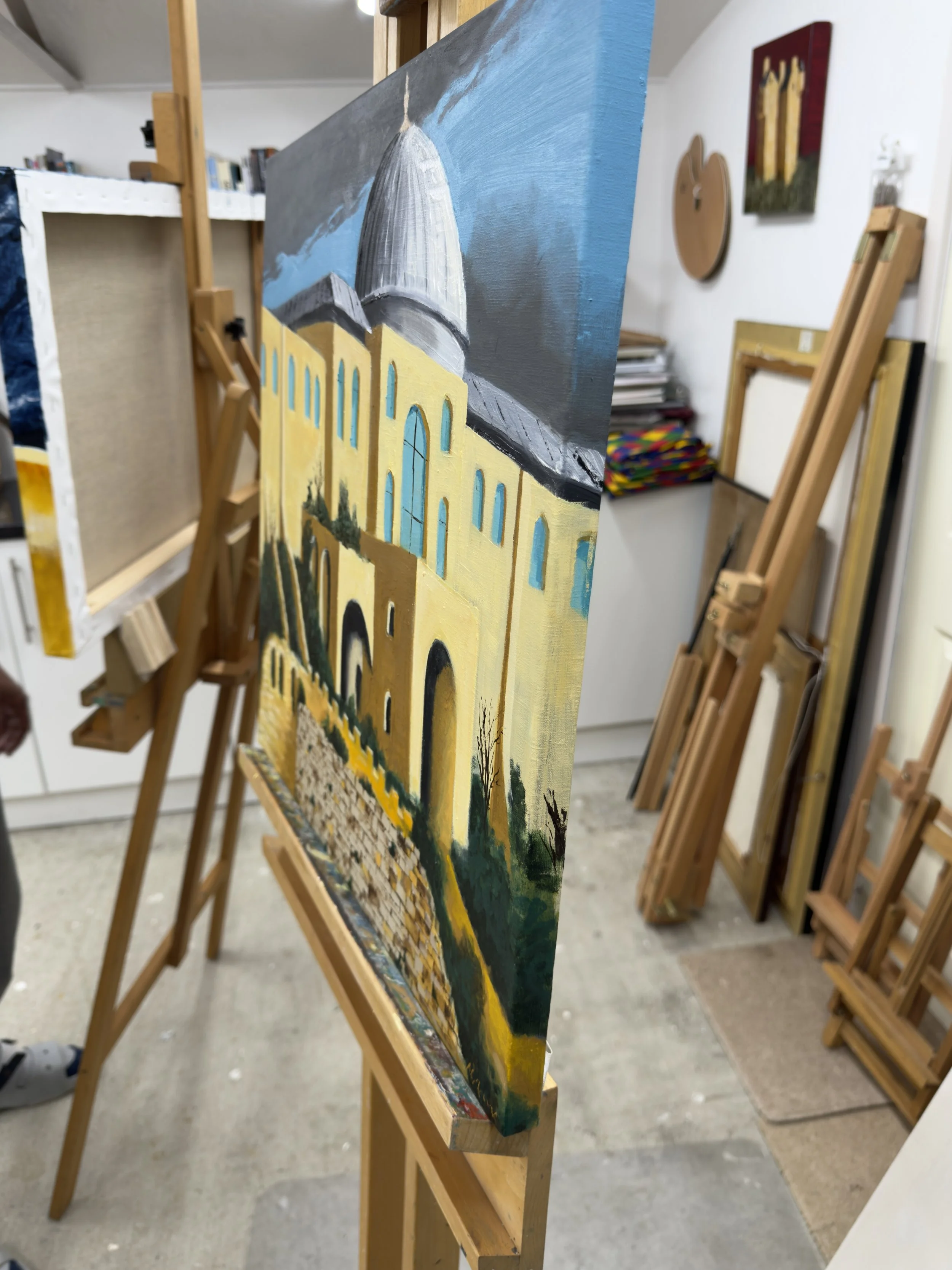A painting of a building with a large dome, set against a sky with clouds, is on an easel in an art studio. The building appears to have yellow walls and arched windows.