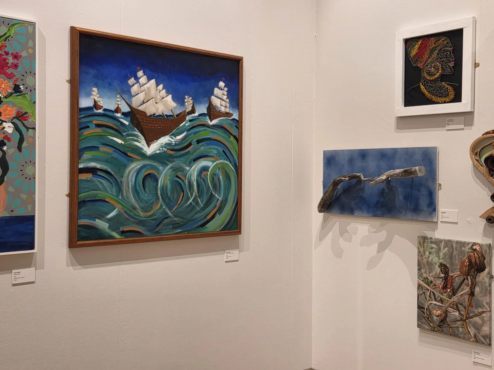 Painting of a sailing ship on turbulent waves, with a dark blue sky background, displayed on a white gallery wall.