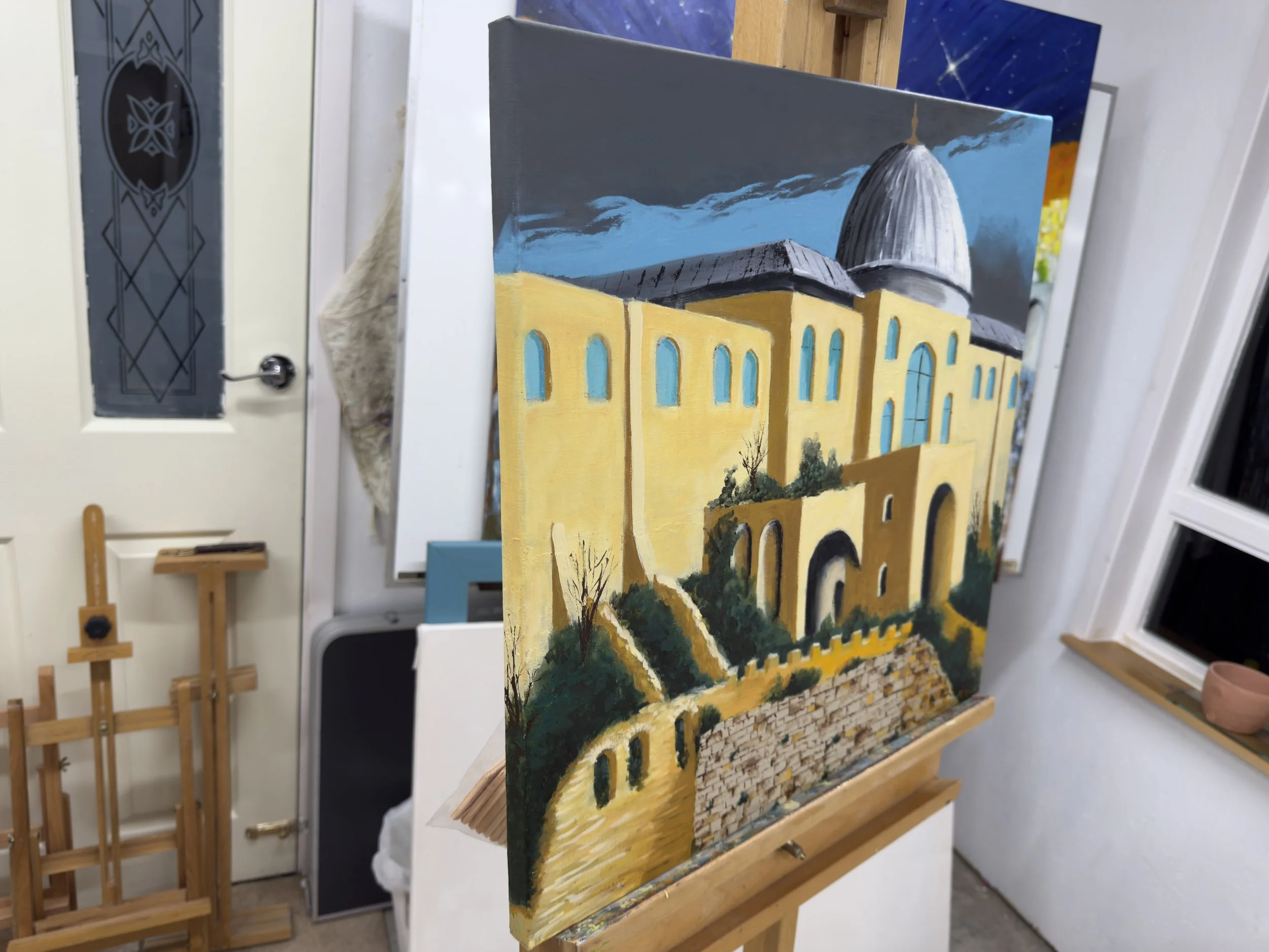 A painting of a yellow building with blue windows and a large gray dome, set against a cloudy sky, displayed on an easel inside an art studio.