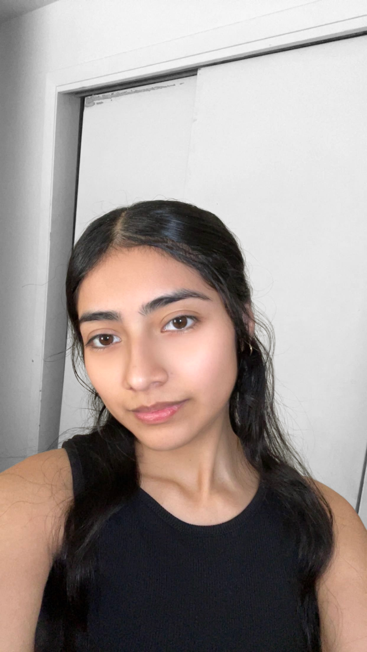 A young woman with dark hair and brown eyes taking a selfie in a room with a white background and a sliding door.