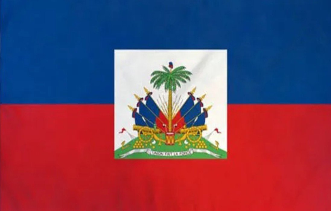 Haitian flag with red and blue horizontal stripes and a white square in the center featuring a coat of arms with a palm tree, cannons, flags, and a banner.