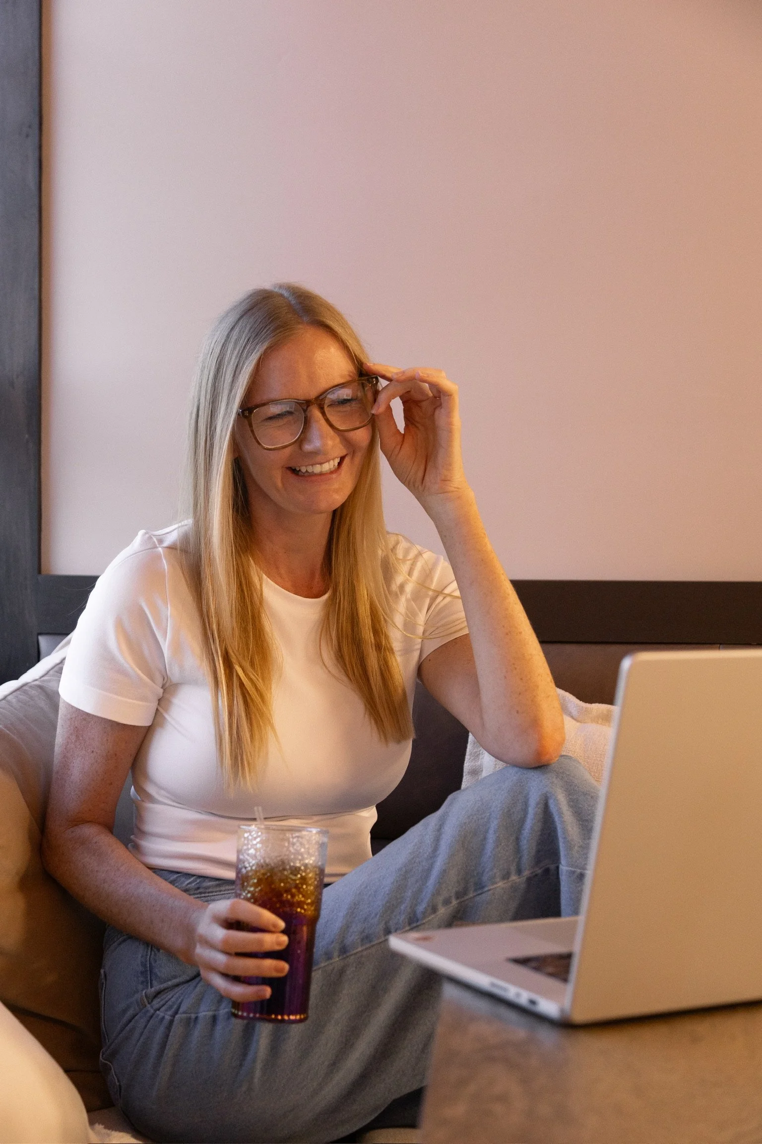 A woman with blonde hair, wearing glasses and a white t-shirt, smiling while sitting on a couch in front of a laptop, holding a glass of soda.