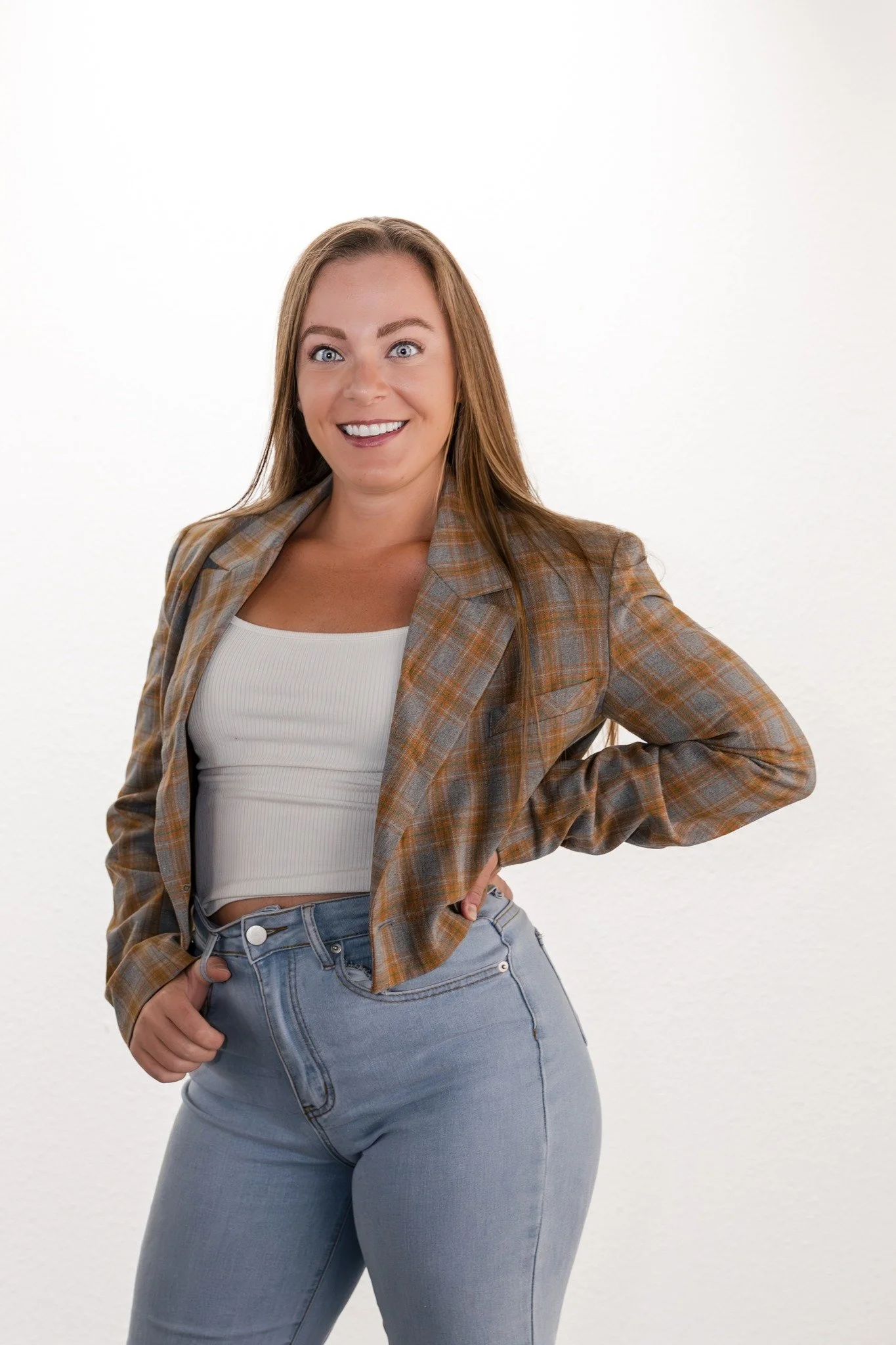 A woman with long brown hair smiling, wearing a brown plaid blazer over a white top and light blue jeans, standing with one hand on her hip against a white background.