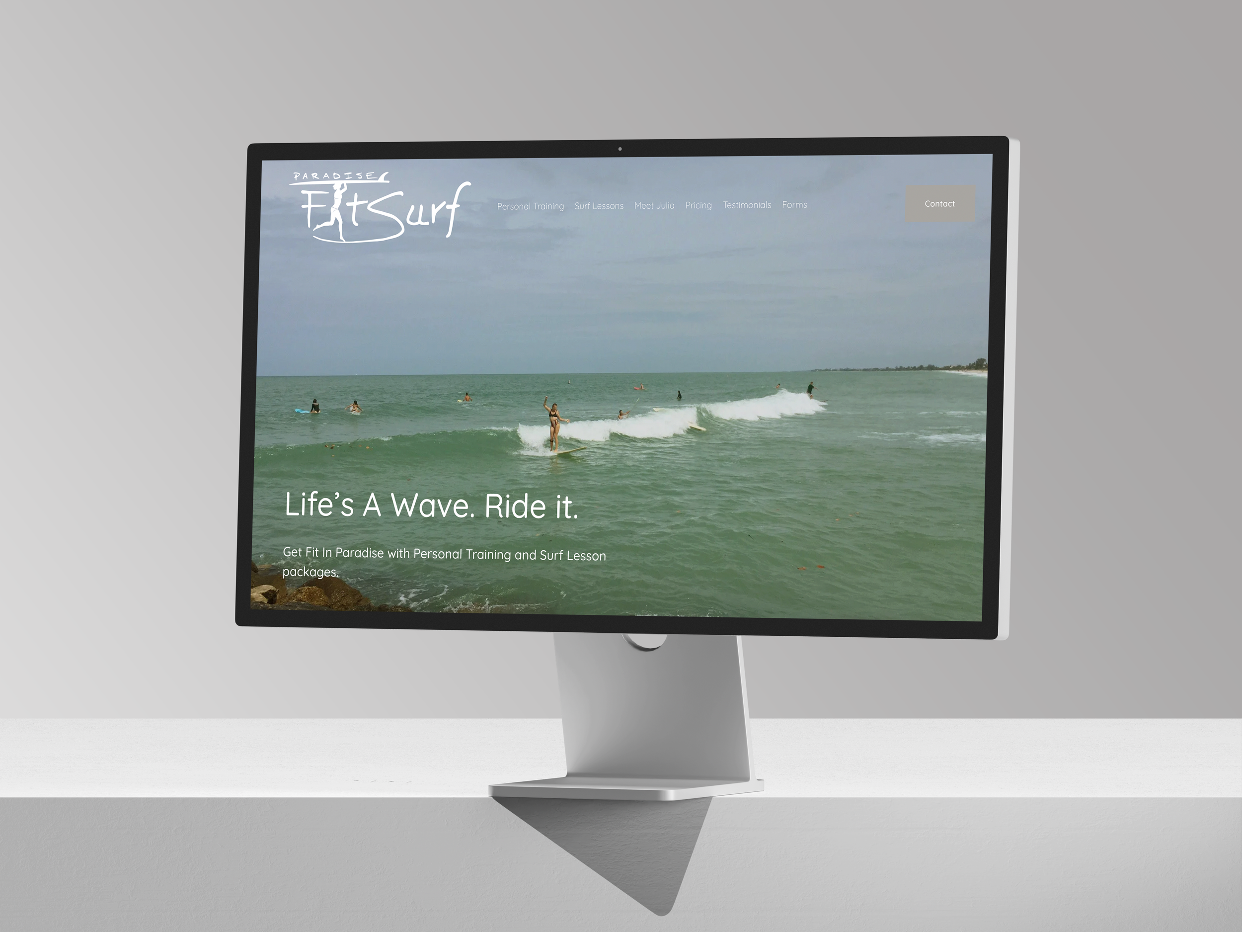Computer monitor displaying a website with a photo of people surfing on the ocean. The website is for a surfing and personal training business called 'Paradise Fit Surf' with a slogan 'Life's A Wave. Ride it.'