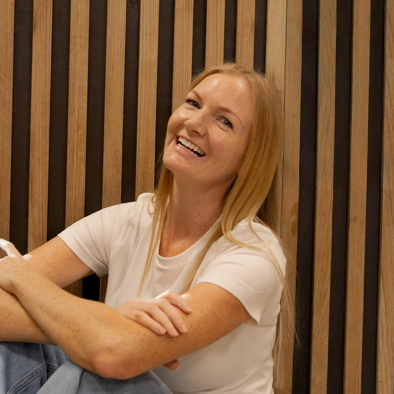 A smiling woman with long blonde hair, wearing a white t-shirt, sitting against a wooden slatted wall.