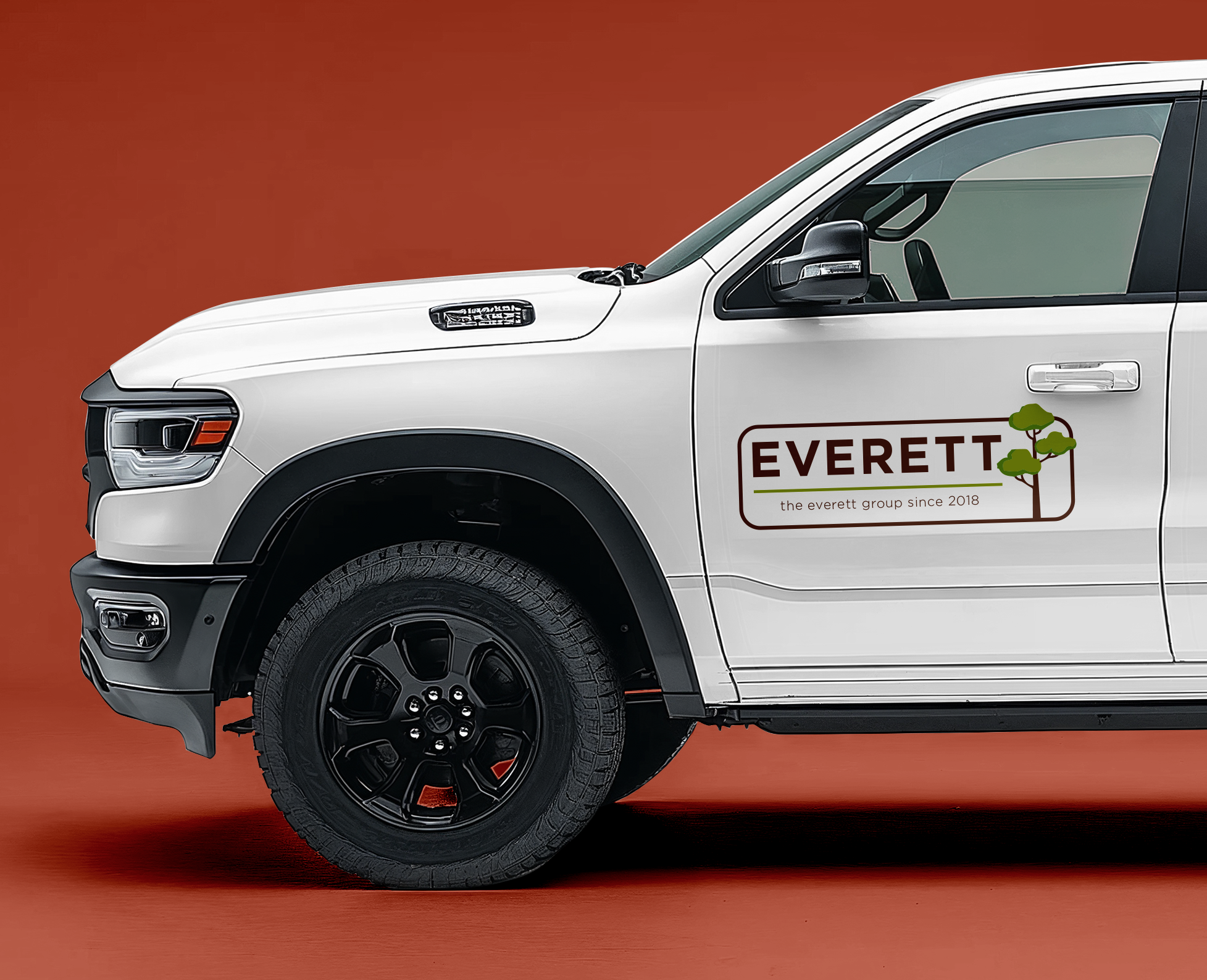 Close-up of a white pickup truck with Everett company logo on the door, featuring trees and text 'the Everett group since 2018', set against a reddish background.