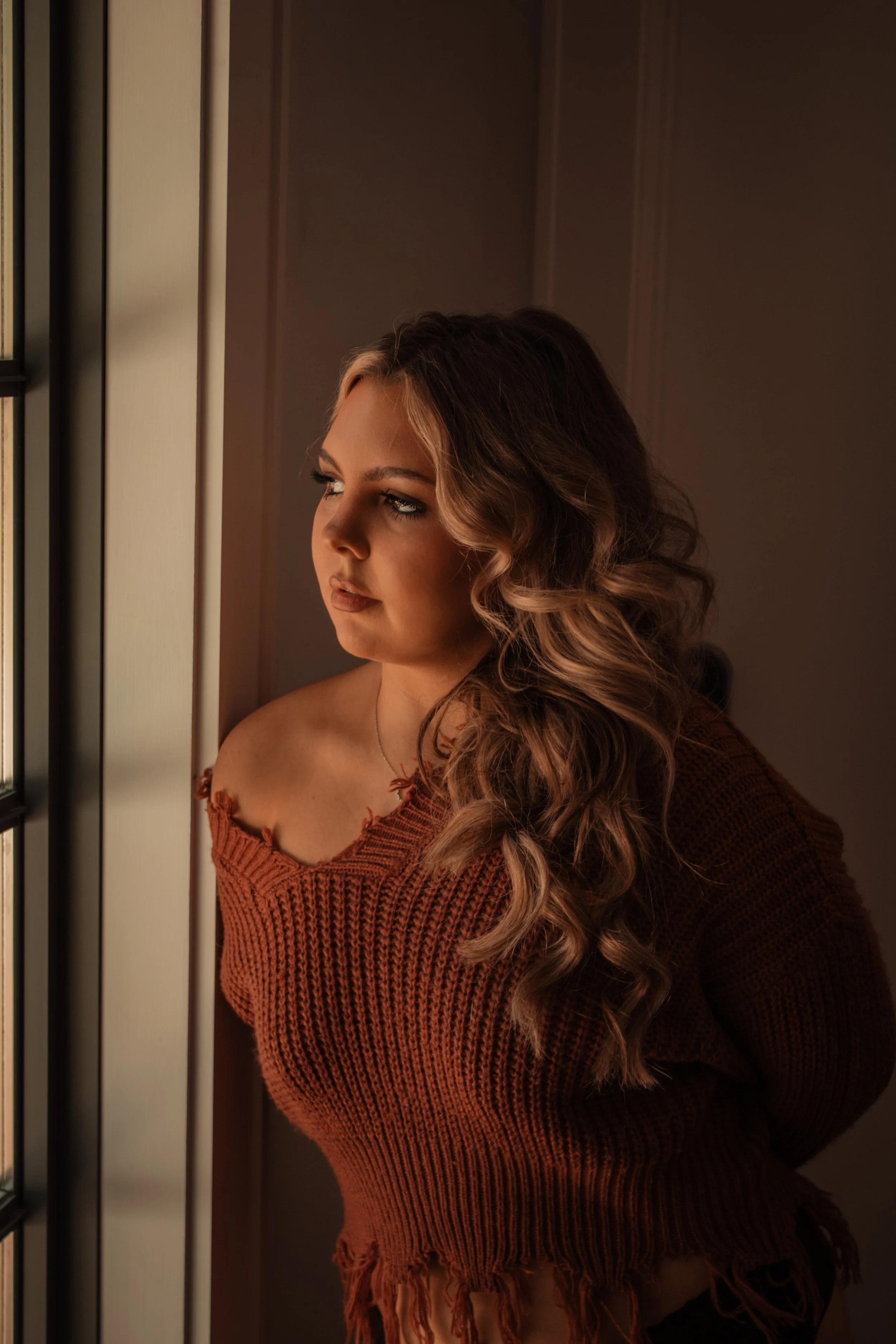 A young woman with long, wavy brown hair stands by a window, gazing outside. She is wearing a burnt orange, off-shoulder, distressed knit sweater.