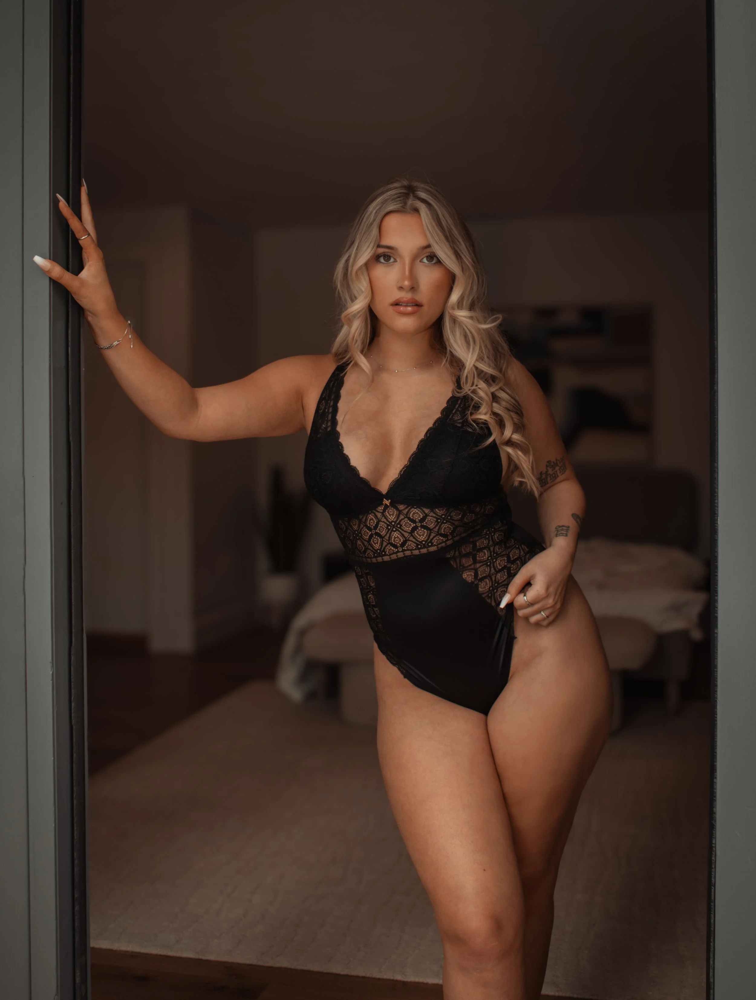A woman with blonde wavy hair in black lace and satin bodysuit posing in a doorway, inside a bedroom.