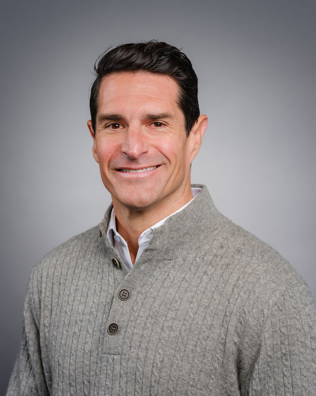 A middle-aged man with dark hair, smiling, wearing a light gray cardigan over a white collared shirt, standing against a gray background.
