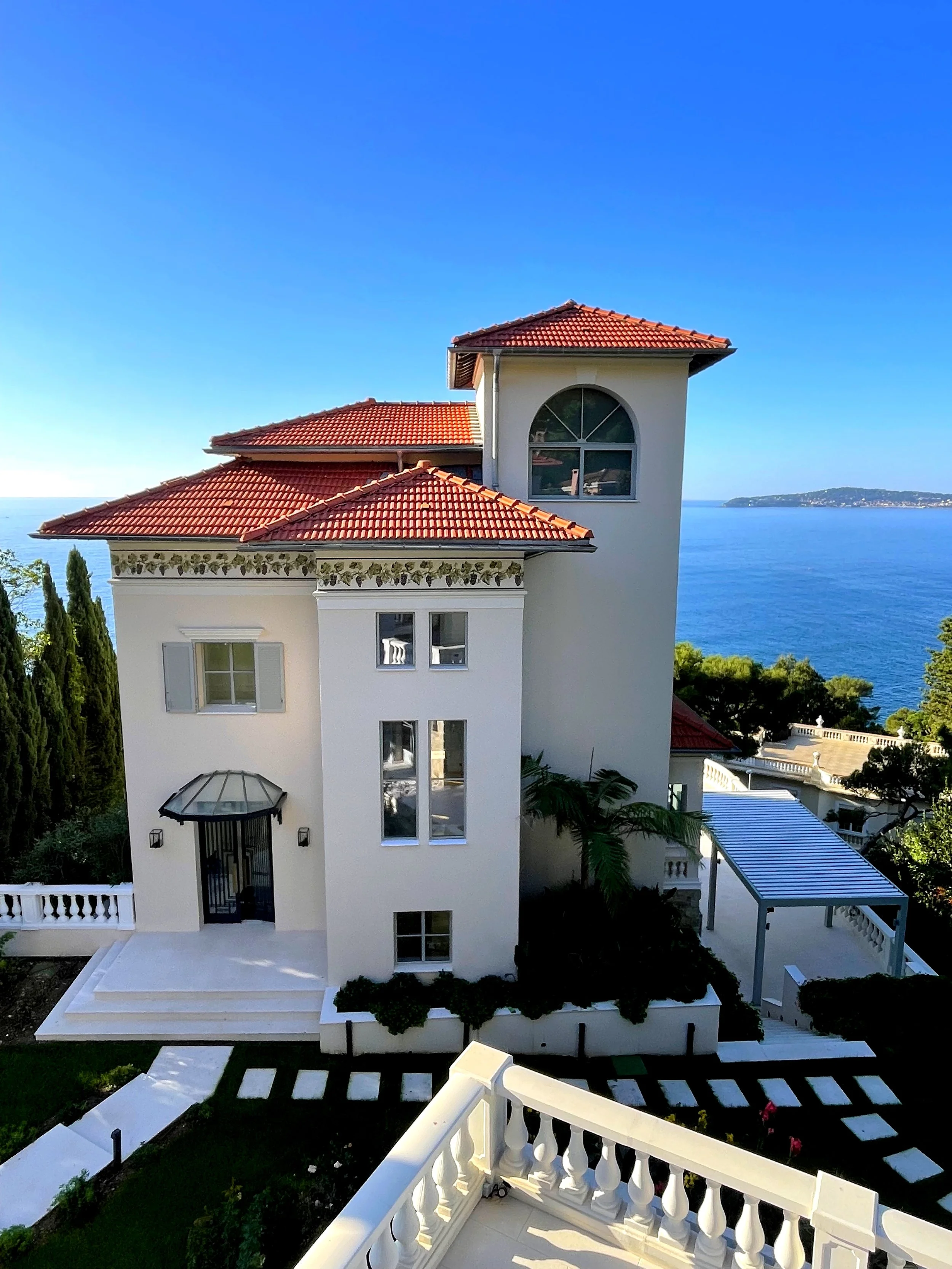 Front view of a white house with red tile roof and multiple levels, overlooking a large body of water in the background, with a balcony railing in the foreground.