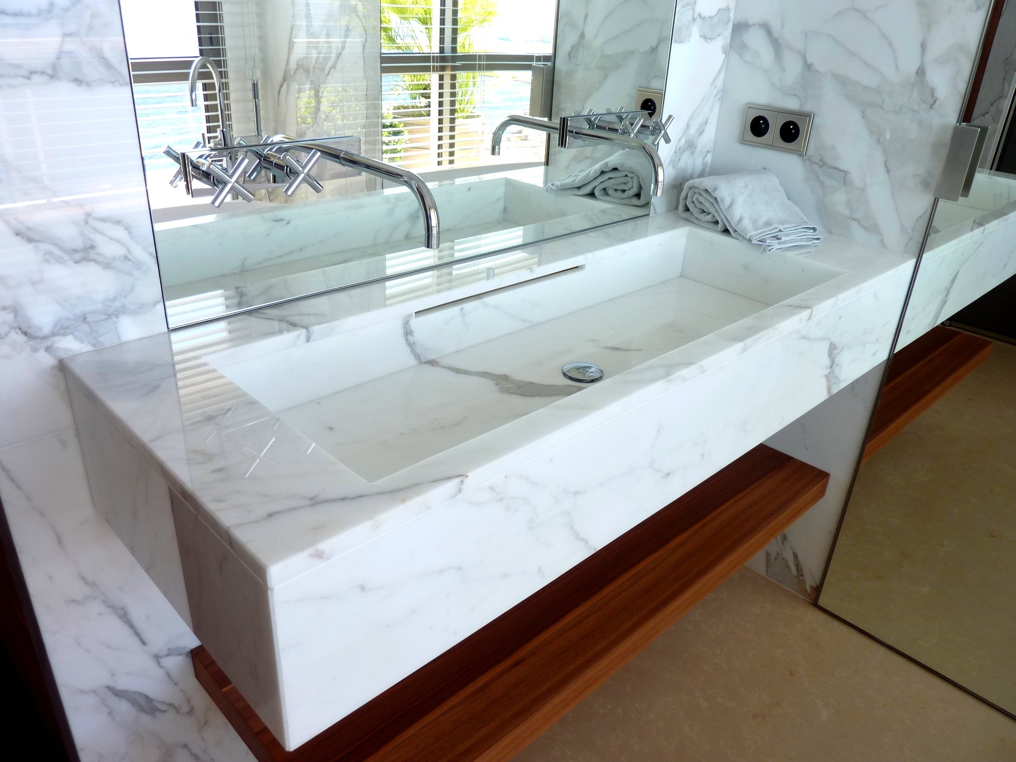 White marble bathroom sink with two chrome faucets and towels near electrical outlets.