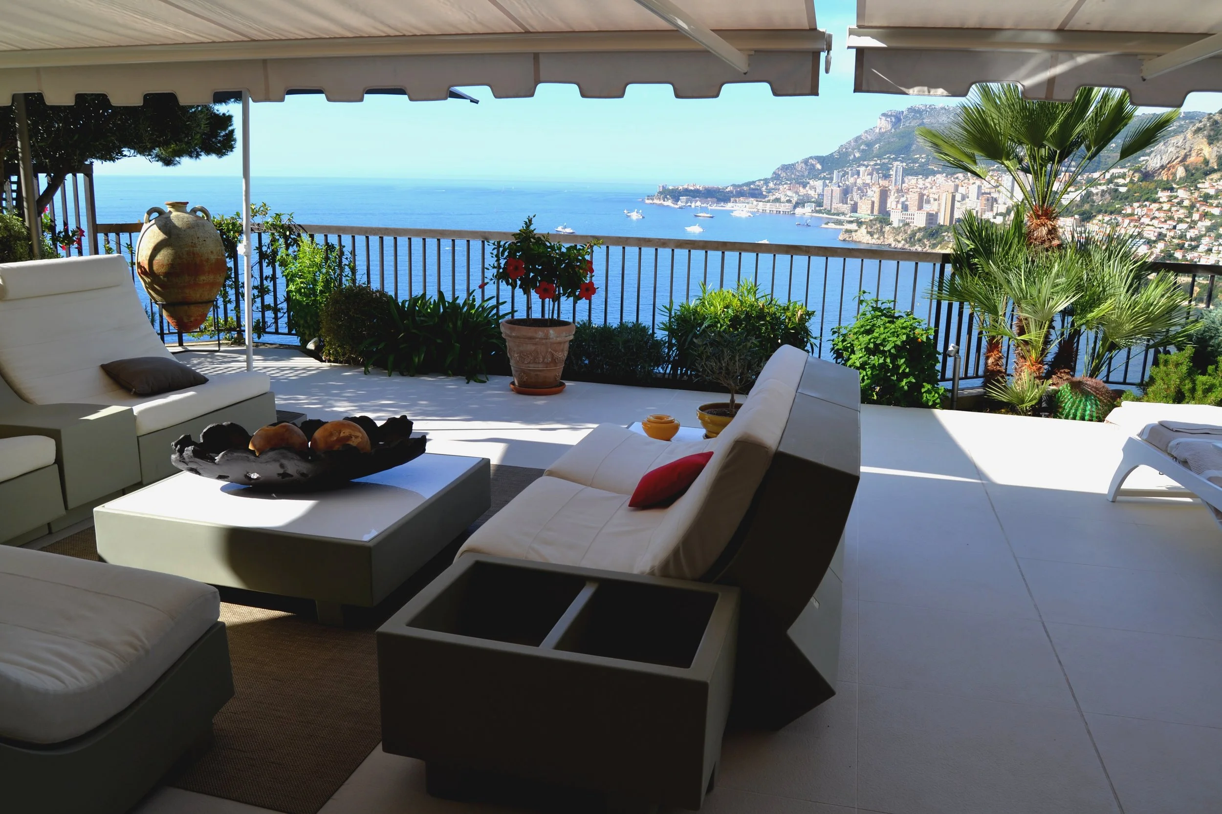 Sea view from a terrace with white outdoor furniture, potted plants, and a railing overlooking the ocean, city, and mountains in the distance.