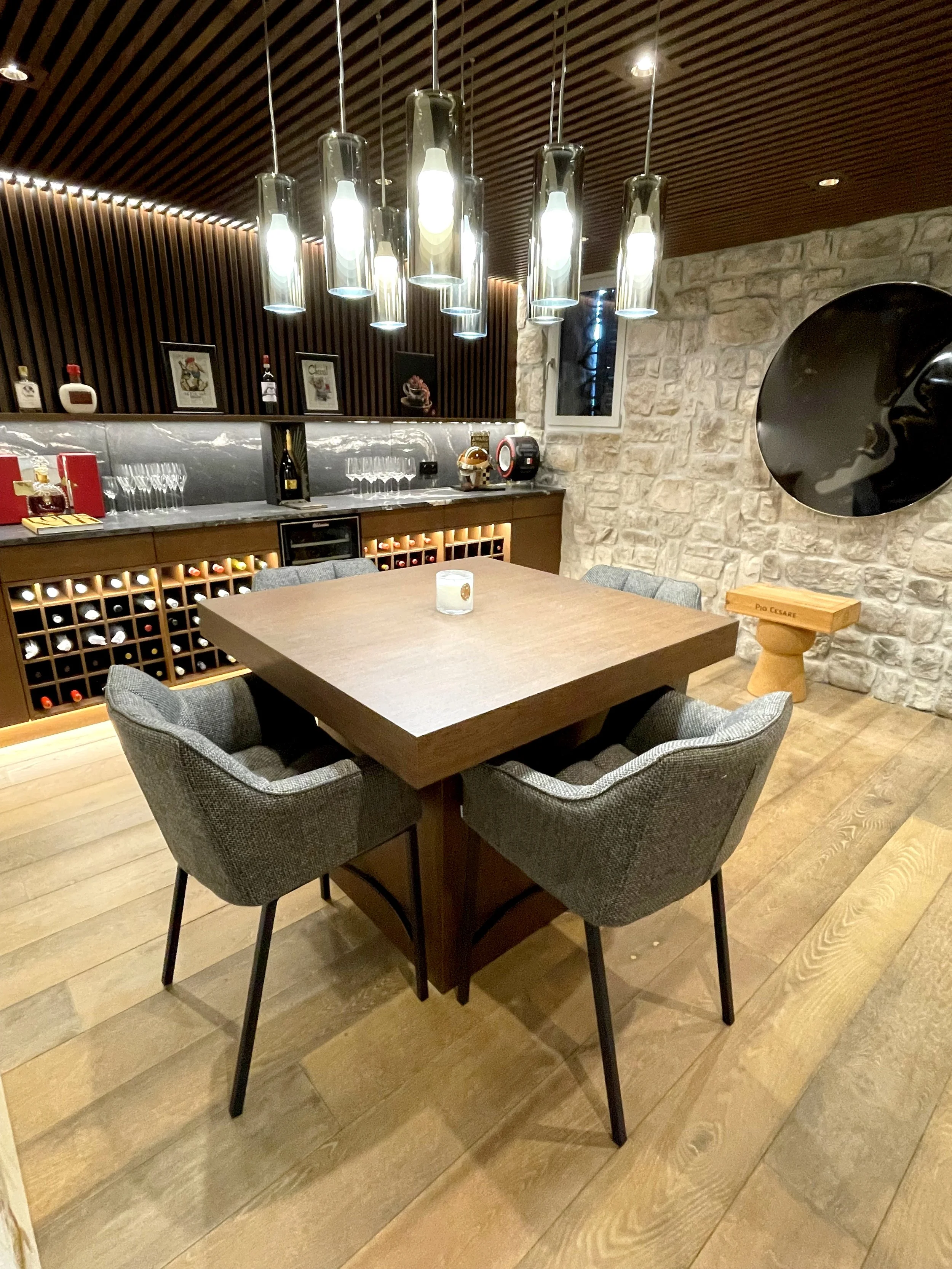 A cozy dining room with a square wooden table surrounded by four gray upholstered chairs. A candle sits on the table. Behind the table, a wine rack and bar area with glasses, bottles, and a wine cooler are visible. The room features a stone wall, modern pendant lights hanging from the wooden slatted ceiling, and decorative pictures on the wall.