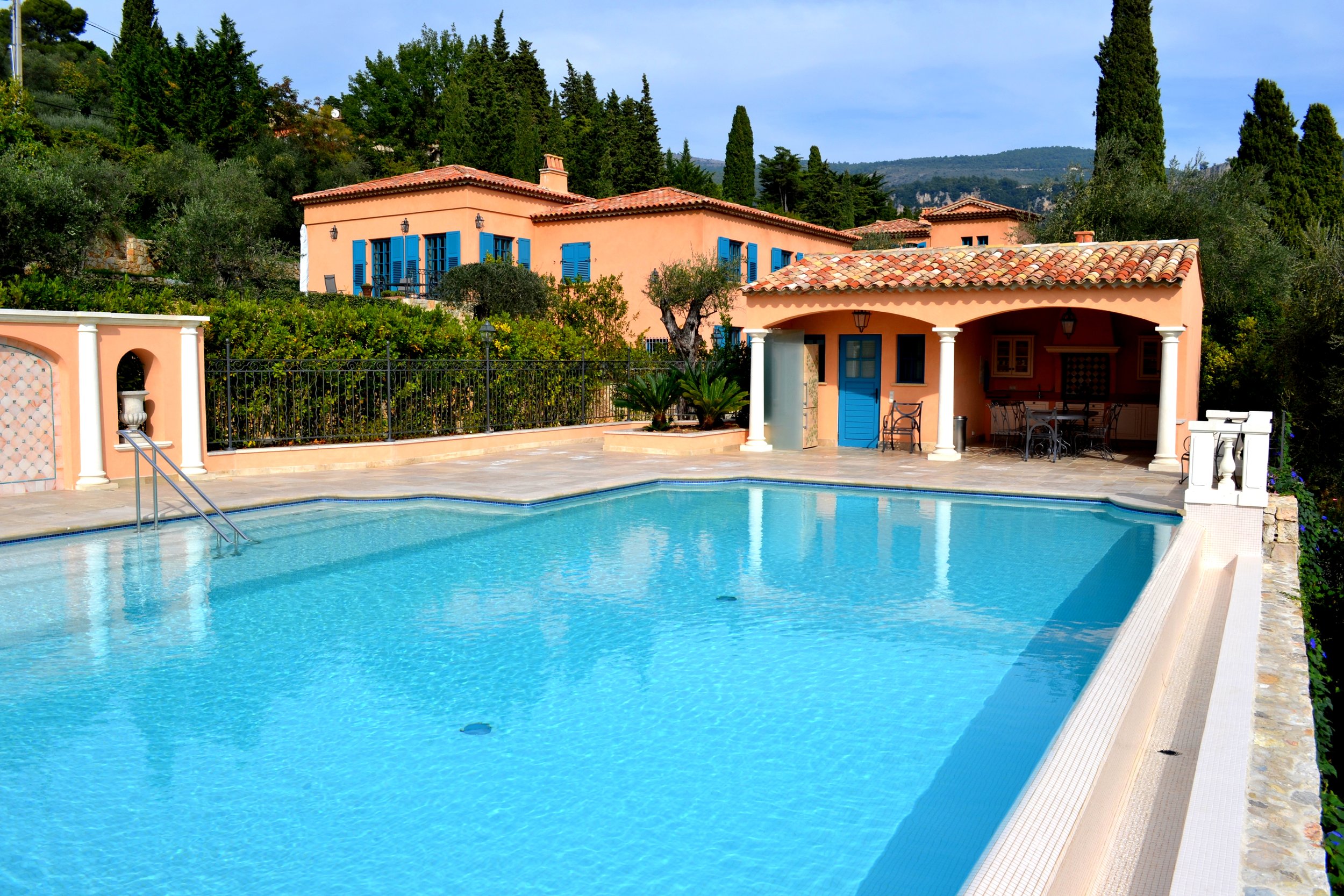 A swimming pool in front of a Mediterranean-style villa with pink walls, blue shutters, and tiled roof amid green trees and hills.