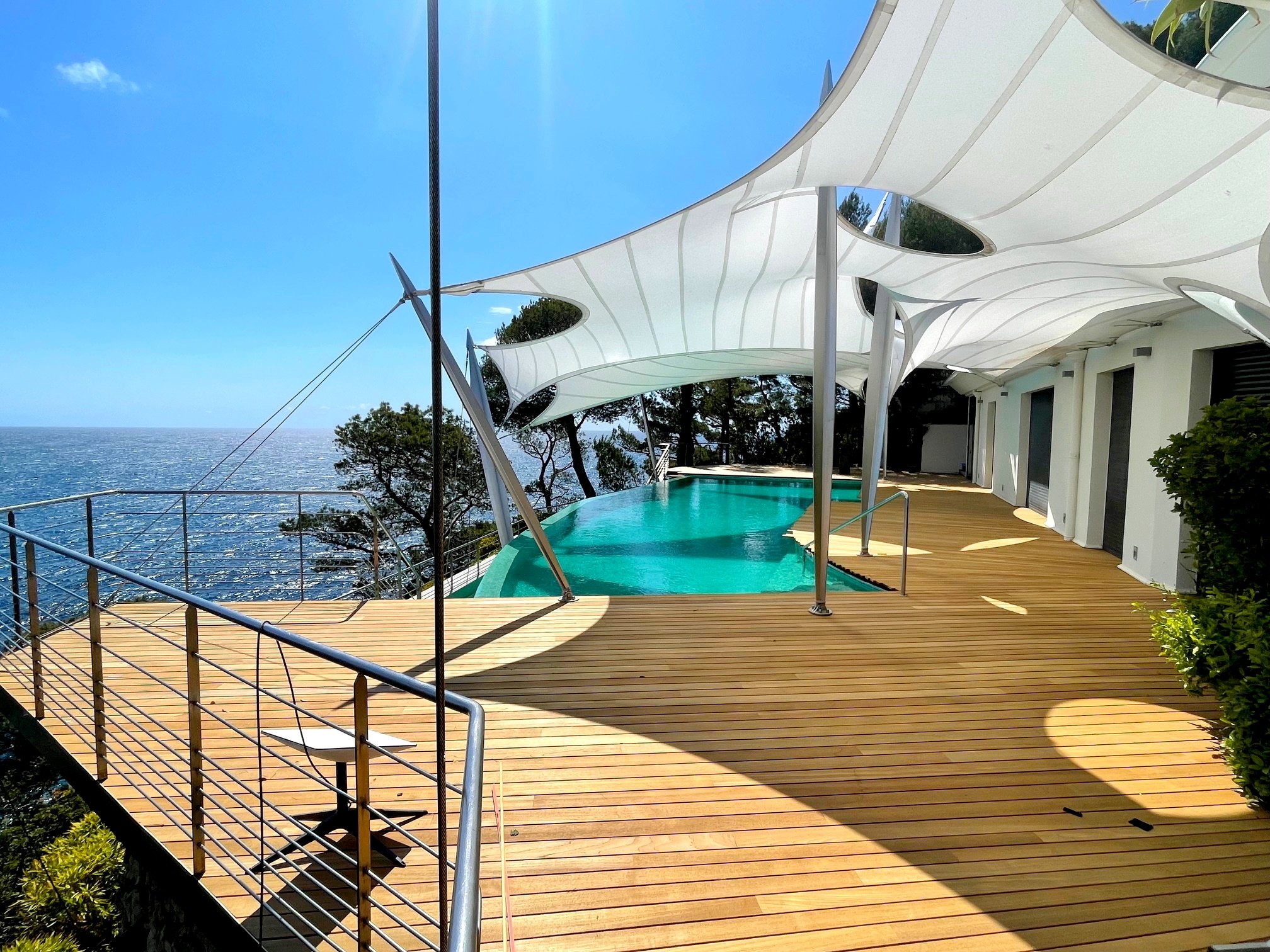 Modern house with a deck overlooking the ocean, featuring an infinity pool, white canopy shades, and greenery.